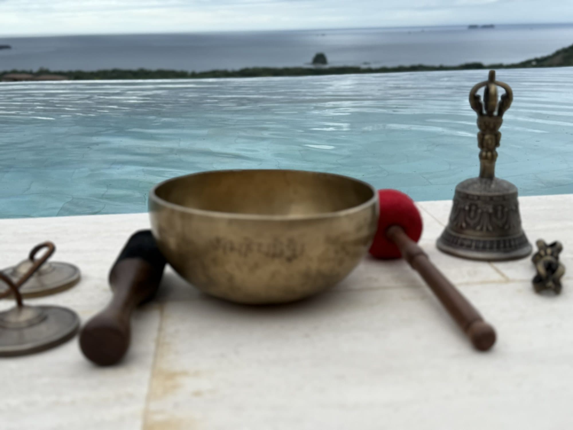 A singing bowl, bell, and tingsha cymbals at the edge of a pool overlooking the water